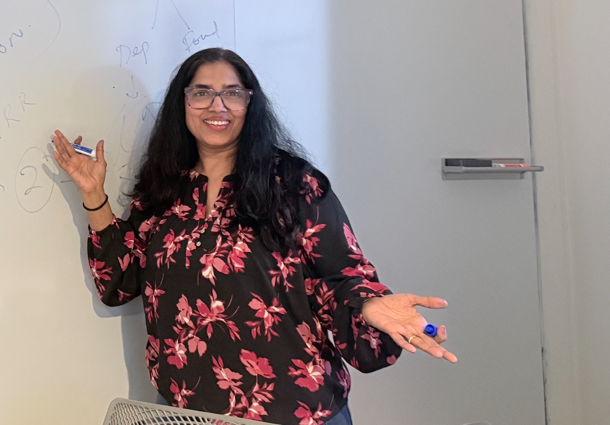 Suneeta standing at a whiteboard with a marker in each hand, arms open, smiling.