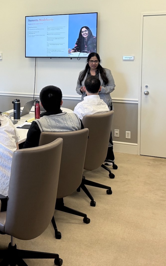 Suneeta speaking beside an opening slide while attendees listen from the conference table.